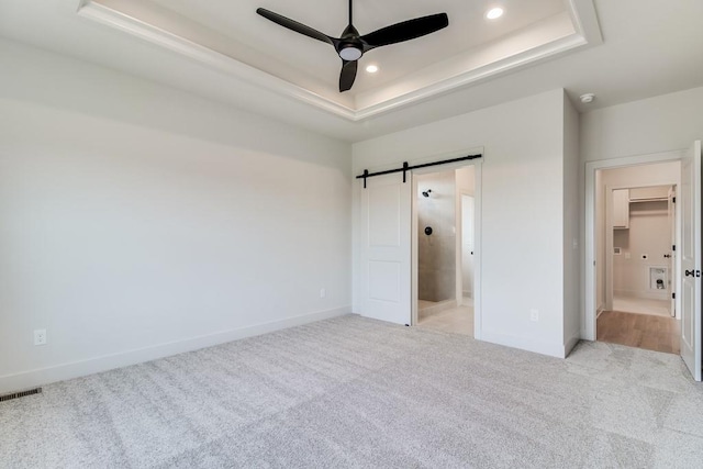 unfurnished bedroom with ensuite bath, ceiling fan, a barn door, a tray ceiling, and light carpet