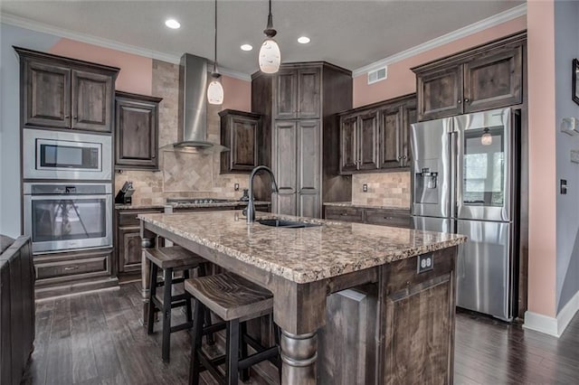 kitchen with appliances with stainless steel finishes, dark brown cabinets, wall chimney exhaust hood, an island with sink, and sink