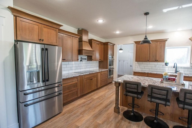 kitchen featuring custom range hood, light wood-type flooring, appliances with stainless steel finishes, decorative backsplash, and decorative light fixtures