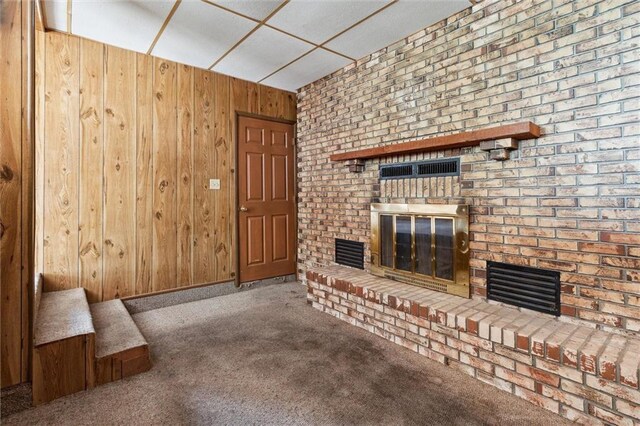 unfurnished living room featuring wooden walls, a drop ceiling, a brick fireplace, and carpet floors