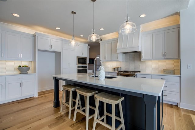 kitchen featuring light wood-type flooring, appliances with stainless steel finishes, pendant lighting, a kitchen island with sink, and white cabinets