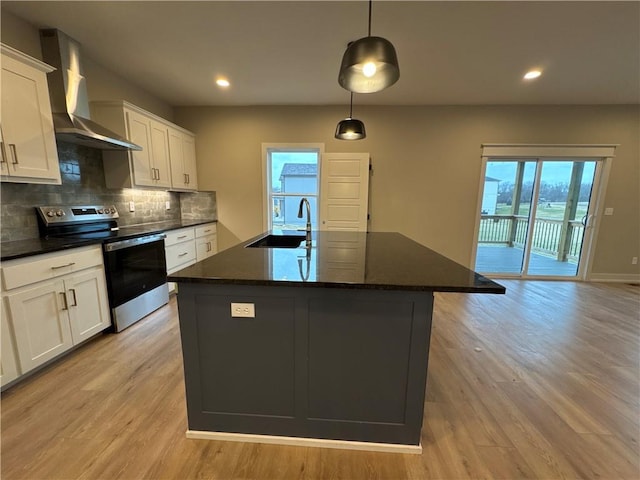 kitchen featuring wall chimney range hood, sink, stainless steel range with electric stovetop, a kitchen island with sink, and decorative light fixtures