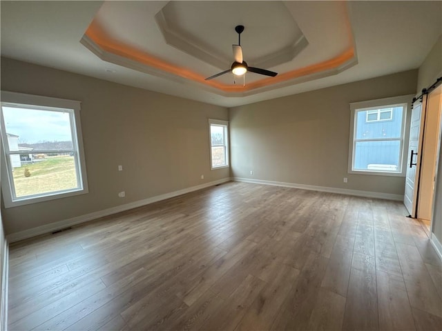 unfurnished room with a tray ceiling, a barn door, ceiling fan, and light wood-type flooring