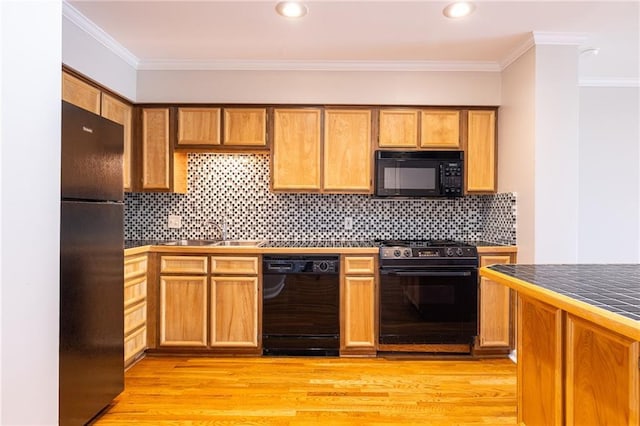 kitchen with a sink, light wood-style floors, tile counters, black appliances, and crown molding