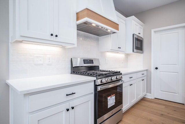 kitchen with tasteful backsplash, white cabinets, custom exhaust hood, stainless steel appliances, and light wood-type flooring