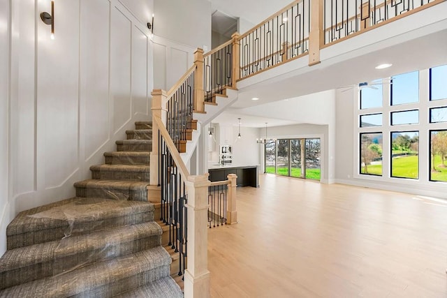 staircase featuring a decorative wall, a towering ceiling, ceiling fan with notable chandelier, and wood finished floors