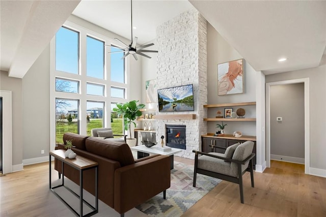 living area with a high ceiling, a stone fireplace, light wood-type flooring, baseboards, and ceiling fan