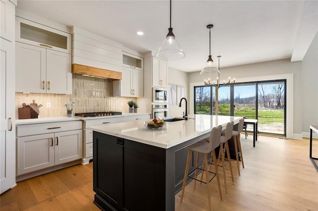 kitchen featuring light wood finished floors, a kitchen island with sink, appliances with stainless steel finishes, a sink, and backsplash