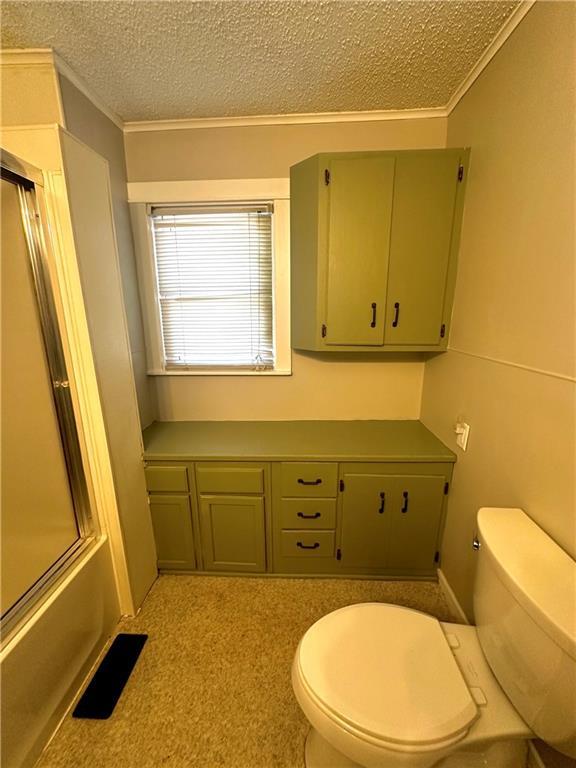 bathroom with a textured ceiling, bath / shower combo with glass door, crown molding, and toilet