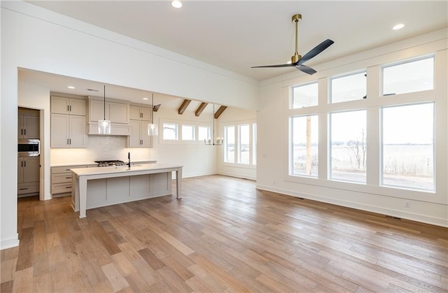 kitchen with tasteful backsplash, stainless steel microwave, a kitchen island with sink, pendant lighting, and light hardwood / wood-style floors