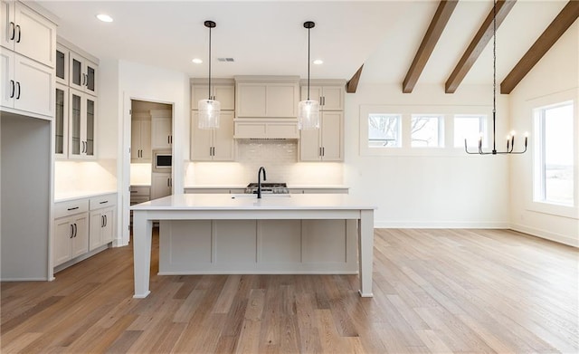 kitchen featuring tasteful backsplash, hanging light fixtures, a kitchen island with sink, beamed ceiling, and light hardwood / wood-style floors