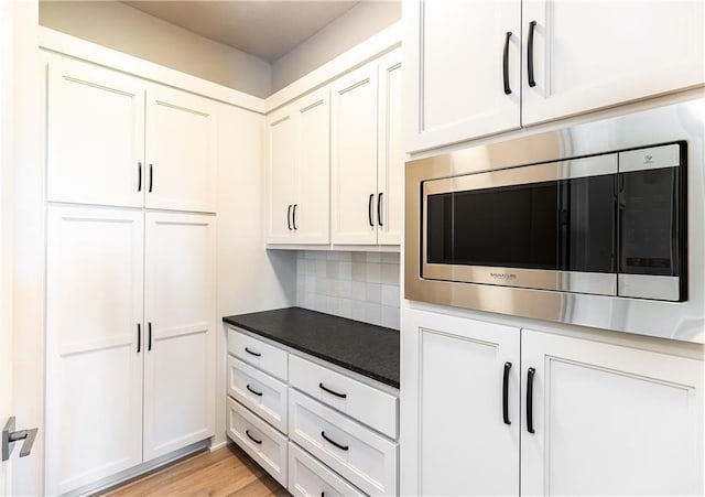 kitchen featuring light wood finished floors, white cabinets, decorative backsplash, dark countertops, and stainless steel microwave