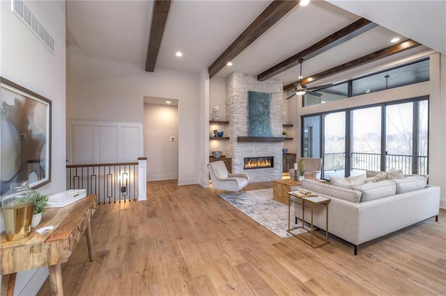 living room with beamed ceiling, ceiling fan, a stone fireplace, and light wood-type flooring