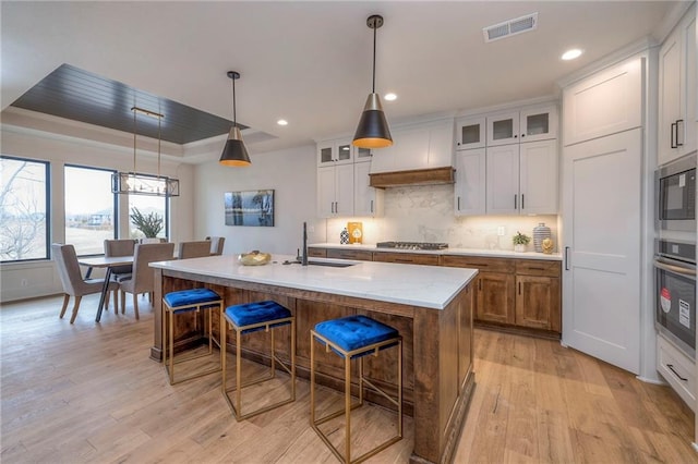 kitchen with white cabinetry, an island with sink, hanging light fixtures, a raised ceiling, and light wood-type flooring