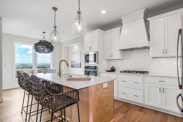 kitchen with premium range hood, pendant lighting, white cabinetry, an island with sink, and light hardwood / wood-style flooring