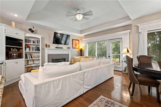 living area with dark wood-style floors, a tray ceiling, a tiled fireplace, and a ceiling fan