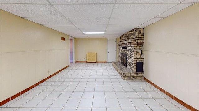 basement featuring light tile patterned floors, a brick fireplace, and a drop ceiling