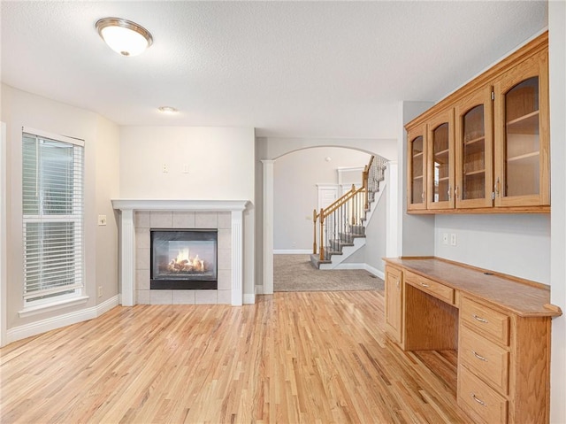 unfurnished living room featuring arched walkways, baseboards, a tile fireplace, light wood-style flooring, and stairs