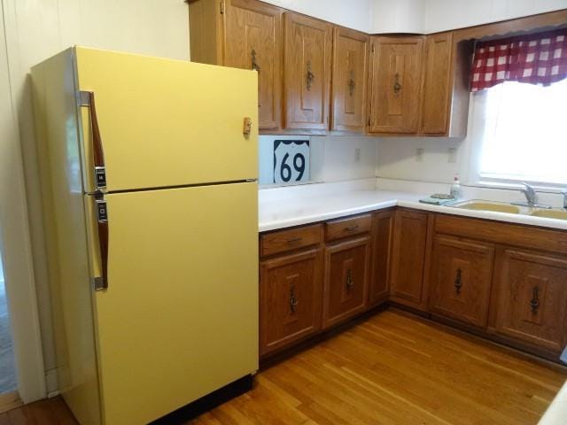 kitchen with brown cabinetry, wood finished floors, freestanding refrigerator, light countertops, and a sink