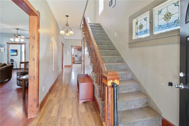 stairway with ceiling fan with notable chandelier, plenty of natural light, wood finished floors, and baseboards