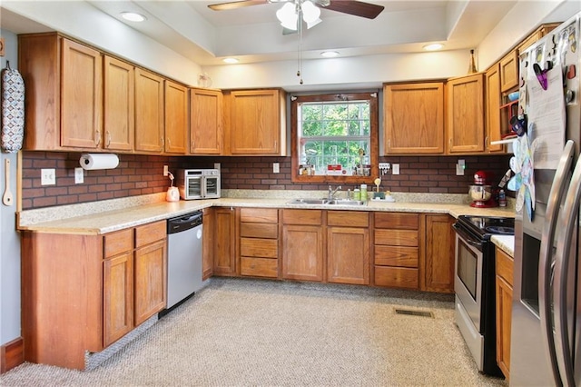 kitchen with a tray ceiling, backsplash, appliances with stainless steel finishes, brown cabinetry, and a sink