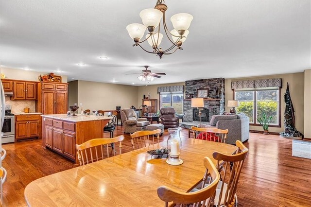 dining space featuring a stone fireplace, ceiling fan with notable chandelier, dark hardwood / wood-style floors, and a healthy amount of sunlight