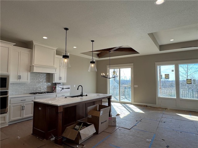 kitchen with decorative backsplash, a raised ceiling, sink, a center island with sink, and white cabinetry