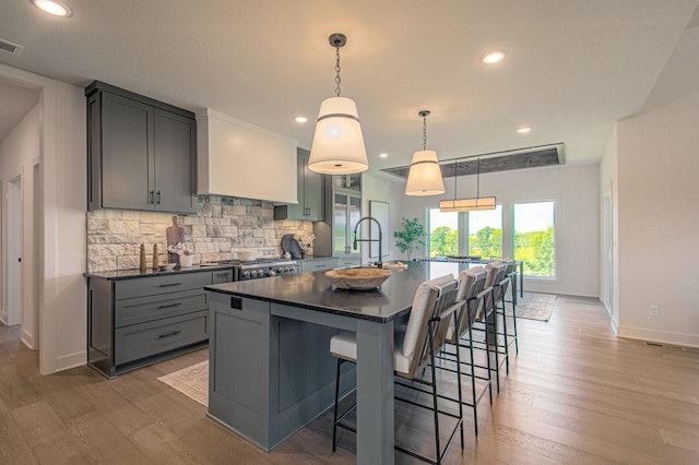 kitchen with decorative backsplash, dark countertops, an island with sink, custom exhaust hood, and gray cabinetry