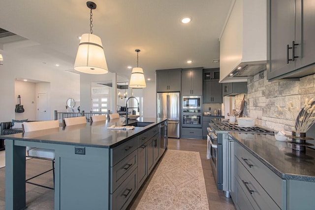 kitchen with gray cabinetry, stainless steel appliances, a sink, wall chimney range hood, and dark countertops