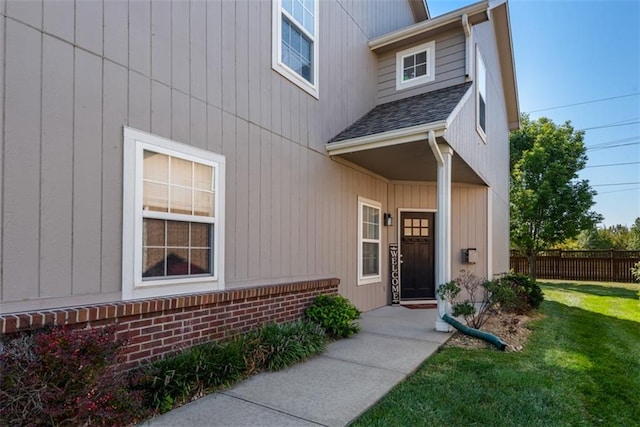 doorway to property with brick siding, a lawn, roof with shingles, and fence