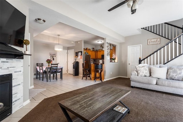 living area featuring baseboards, stairs, a stone fireplace, light tile patterned flooring, and a ceiling fan