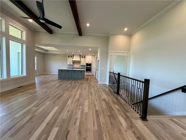 unfurnished living room featuring light wood-style flooring, beamed ceiling, baseboards, a ceiling fan, and crown molding