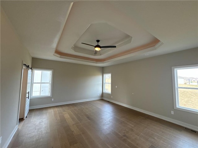 empty room with wood-type flooring, a barn door, ceiling fan, and a tray ceiling