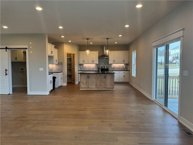 kitchen with an island with sink, wall chimney range hood, white cabinets, and decorative light fixtures