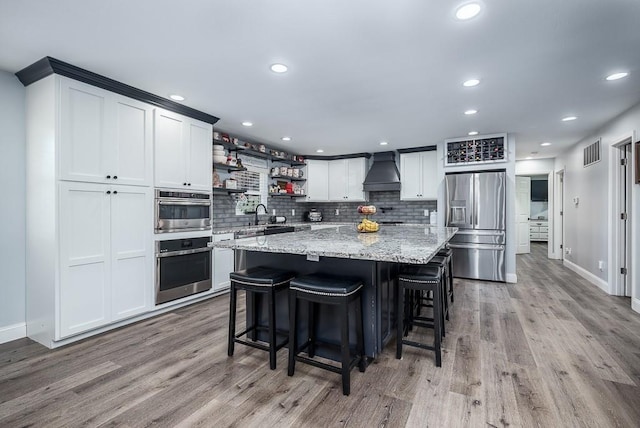 kitchen with stainless steel appliances, light stone counters, custom range hood, white cabinets, and decorative backsplash