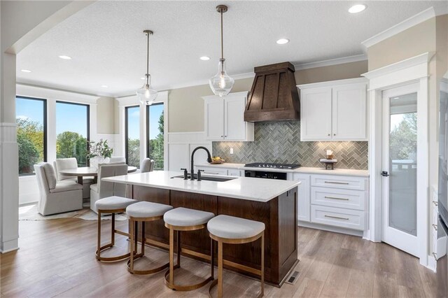 kitchen with light wood-type flooring, custom range hood, sink, and a center island with sink