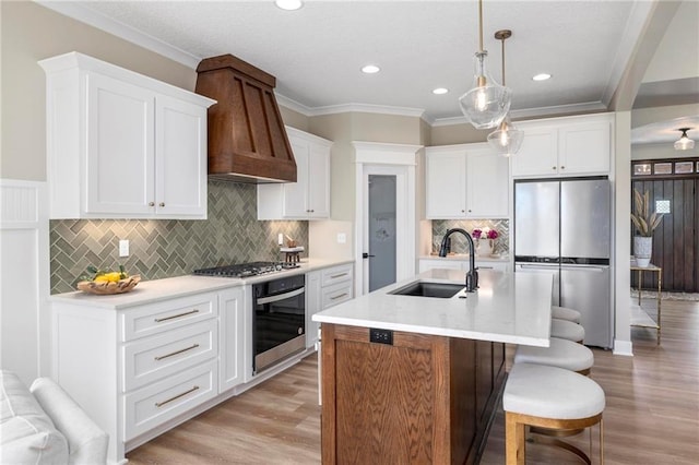 kitchen featuring premium range hood, appliances with stainless steel finishes, an island with sink, sink, and light wood-type flooring