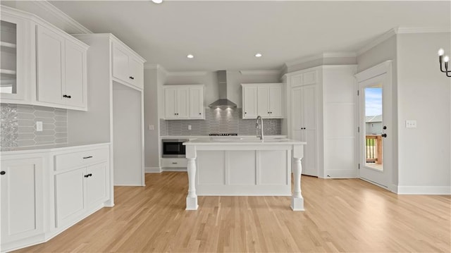 kitchen with built in microwave, white cabinetry, decorative backsplash, a kitchen island with sink, and wall chimney range hood