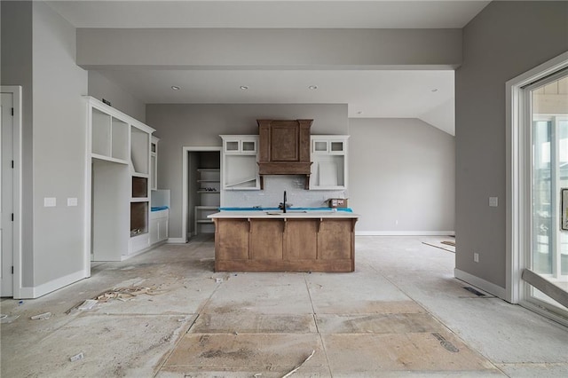 kitchen with a kitchen island with sink, a sink, baseboards, light countertops, and glass insert cabinets