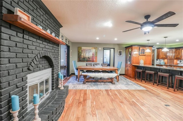 dining space featuring a textured ceiling, a ceiling fan, baseboards, light wood-type flooring, and a brick fireplace