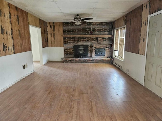 unfurnished living room featuring hardwood / wood-style flooring, wooden walls, a brick fireplace, a baseboard radiator, and ceiling fan