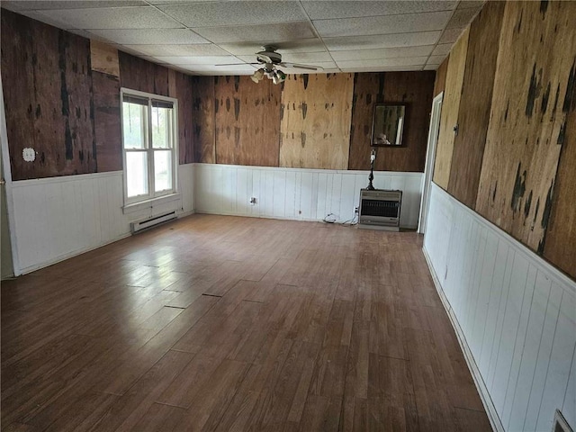 unfurnished room featuring wooden walls, a drop ceiling, wood-type flooring, a baseboard radiator, and ceiling fan