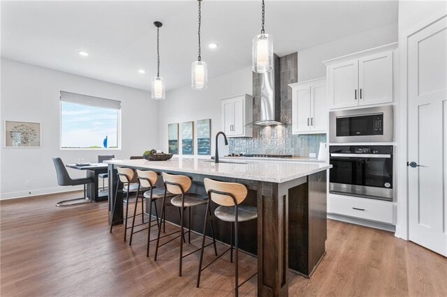 kitchen with an island with sink, sink, stainless steel oven, wall chimney exhaust hood, and black microwave