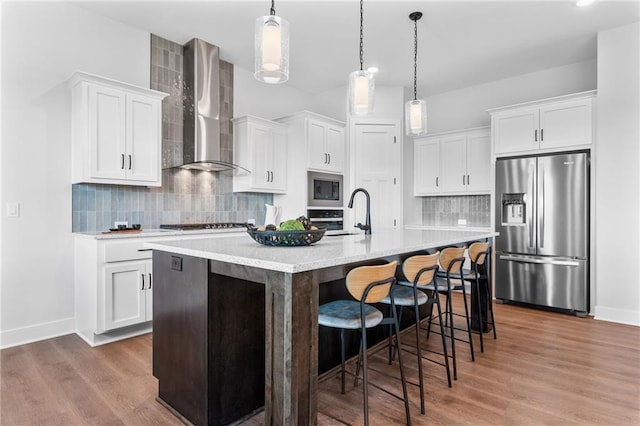 kitchen featuring a kitchen island with sink, wood finished floors, light countertops, appliances with stainless steel finishes, and wall chimney exhaust hood