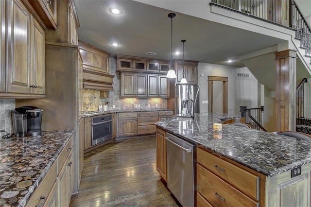 kitchen featuring a kitchen island with sink, decorative backsplash, dark stone countertops, dark hardwood / wood-style flooring, and stainless steel appliances