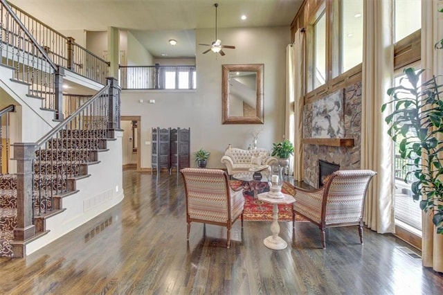 sitting room featuring dark hardwood / wood-style floors, ceiling fan, a stone fireplace, and a towering ceiling