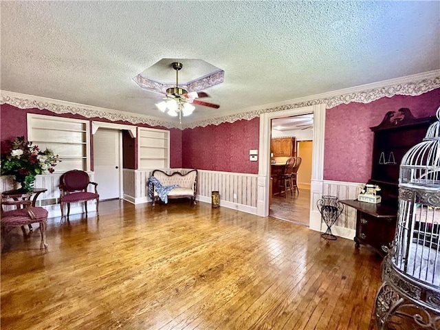 sitting room with ceiling fan, a textured ceiling, a wainscoted wall, wood-type flooring, and crown molding