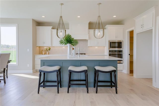 kitchen with hanging light fixtures, light hardwood / wood-style floors, white cabinetry, an island with sink, and decorative backsplash