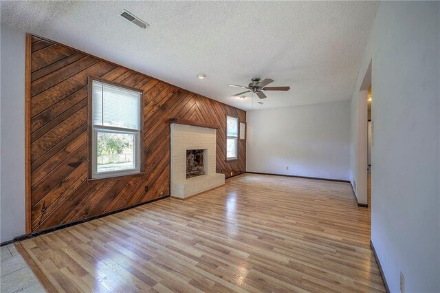 unfurnished living room with a textured ceiling, wood walls, a brick fireplace, ceiling fan, and light wood-type flooring