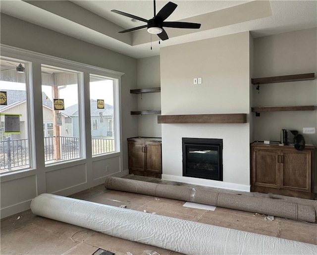 unfurnished living room with a tray ceiling, a glass covered fireplace, a ceiling fan, and baseboards